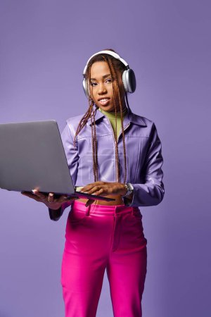 young african american woman with headphones working on laptop remotely on purple background
