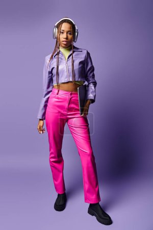 young african american woman with headphones holding laptop and standing on purple background