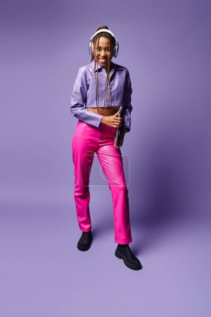 happy african american woman with headphones holding laptop and standing on purple background