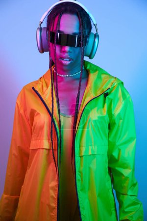 Foto de Joven afroamericana chica en auriculares inalámbricos y chaqueta de pie en el estudio con luces de neón - Imagen libre de derechos