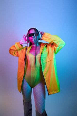 young african american model in wireless headphones and jacket standing in studio with neon lights