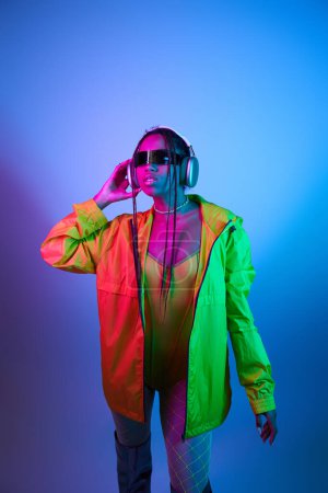 young african american woman in headphones posing in bodysuit and jacket in studio with neon lights