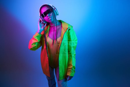 pretty african american woman in headphones posing in bodysuit and jacket in studio with neon lights
