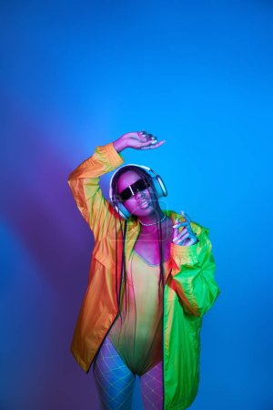 pretty african american girl in headphones posing in bodysuit and jacket in studio with neon lights