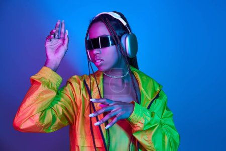 Téléchargez les photos : Afro-américaine dans des écouteurs et des lunettes de soleil élégantes écouter de la musique en studio avec des lumières - en image libre de droit