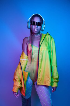 pretty dark-skinned girl in headphones posing in bodysuit and jacket in studio with neon lights