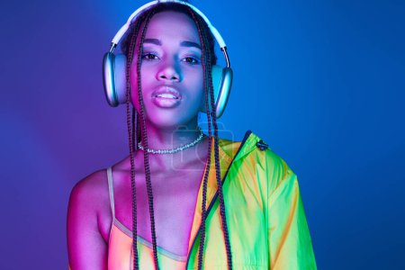 Photo for Beautiful dark-skinned woman in wireless headphones posing in jacket in studio with neon lights - Royalty Free Image