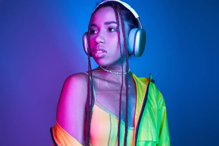 Nachdenkliche dunkelhäutige Frau mit drahtlosen Kopfhörern posiert in Jacke im Studio mit Neonlicht