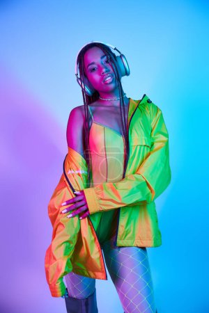 Charmante dunkelhäutige Frau mit drahtlosen Kopfhörern posiert in Jacke im Studio mit Neonlicht