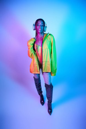 dark-skinned woman in headphones posing in bodysuit and over knee boots in studio with neon lights