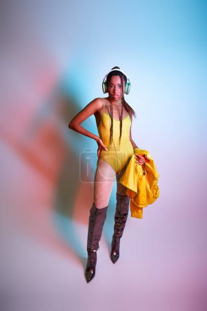 young dark-skinned woman in headphones posing in bodysuit and over knee boots while holding jacket