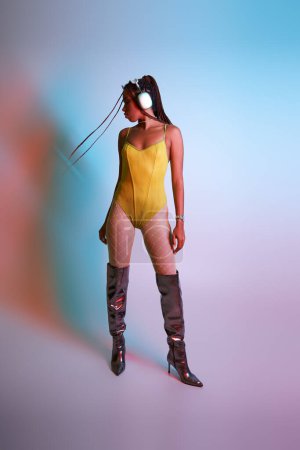 Photo for African american woman in headphones posing in yellow bodysuit and over knee boots, shake head - Royalty Free Image