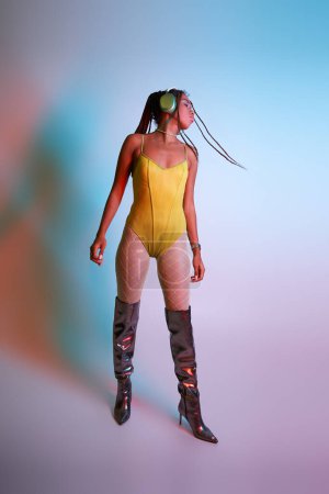 young african american woman in headphones posing in yellow bodysuit and over knee boots, shake head