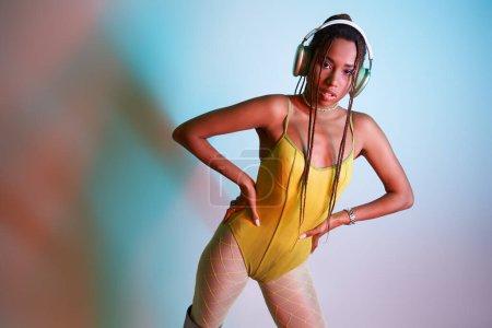 junge Afroamerikanerin mit Kopfhörern posiert im gelben Body mit den Händen auf den Hüften im Studio