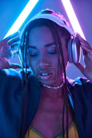 Téléchargez les photos : Jeune fille afro-américaine dans dreadlocks et écouteurs en studio avec lumière fluorescente bleue - en image libre de droit