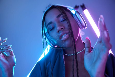 Foto de Joven afroamericana mujer en auriculares disfrutando de la música en estudio con luz fluorescente azul - Imagen libre de derechos