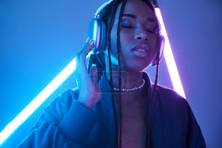 Foto de Encantadora mujer afroamericana en auriculares disfrutando de la música en el estudio con luz fluorescente - Imagen libre de derechos