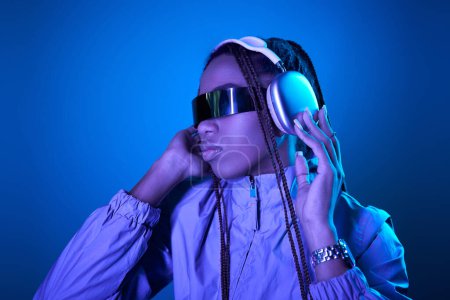 pretty african american woman posing in trendy sunglasses with headphones on blue with neon light