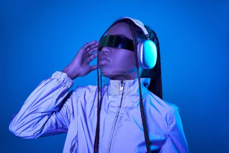 Photo for Pretty dark skinned woman posing in trendy sunglasses with headphones on blue with neon light - Royalty Free Image