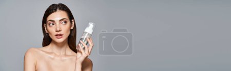Junge kaukasische Frau mit brünetten Haaren sieht überrascht aus, während sie eine Flasche Gesichtsreiniger im Studio hält, Banner