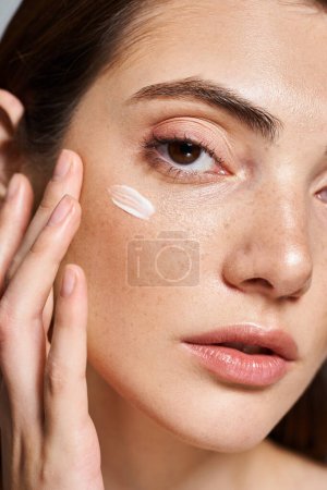 Photo for A young Caucasian woman with brunette hair applying cream to her face, enhancing her natural beauty in a studio setting. - Royalty Free Image
