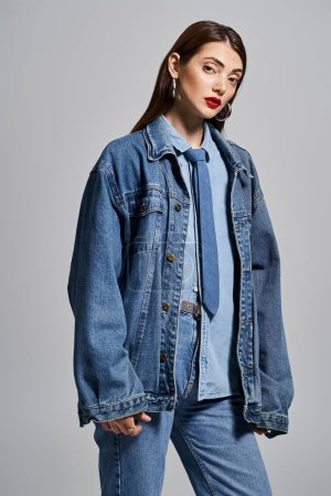 A young Caucasian woman with brunette hair and red lips stylishly wearing a denim jacket and jeans in a studio setting.