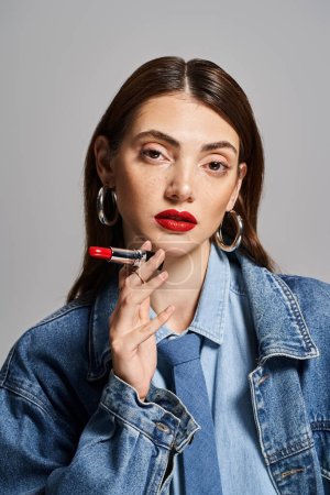 A stylish young woman in a denim jacket holding red lipstick  and looking at camera in studio