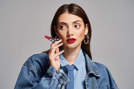 A young Caucasian woman with brunette hair in a denim jacket holding red lipstick