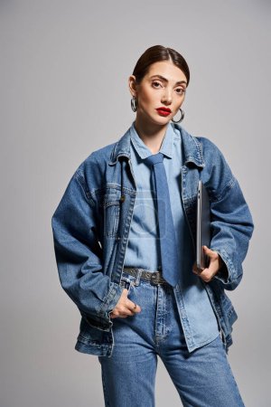 A stylish young Caucasian woman with brunette hair wearing a denim jacket and tie and holding laptop