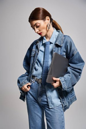 Eine junge Kaukasierin mit brünetten Haaren in Jeansjacke hält selbstbewusst einen Laptop in der Hand.