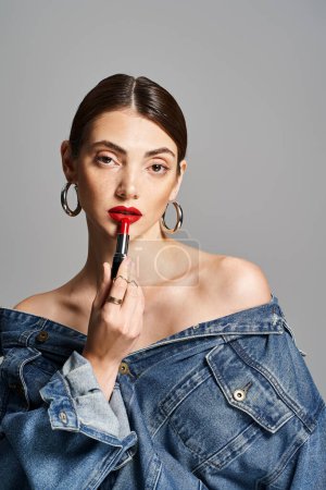 A young Caucasian woman with brunette hair wearing a jean jacket, holding a vibrant red lipstick.