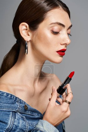 Une jeune femme caucasienne aux cheveux bruns et à la peau propre, portant du rouge à lèvres, tient un tube de rouge à lèvres dans un cadre studio.