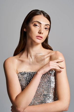 A young Caucasian woman with brunette hair poses in a stunning silver dress, exuding elegance and confidence.