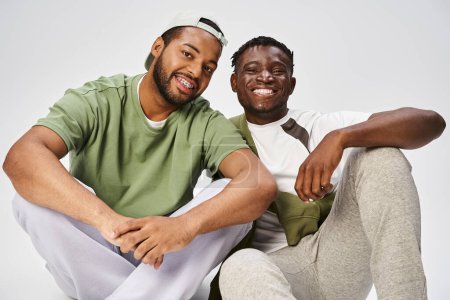 fröhliche Junifeier, junger afrikanisch-amerikanischer Mann sitzt mit männlichem Freund auf grauem Hintergrund