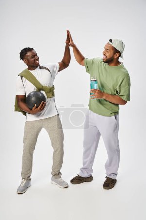 happy african american male friends holding ball and water while giving high five on grey backdrop