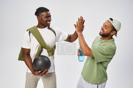 fröhliche afrikanisch-amerikanische männliche Freunde halten Ball und Wasser in der Hand und geben High Five auf grauem Hintergrund