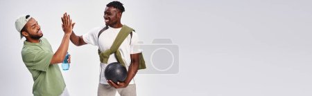 Téléchargez les photos : Joyeux amis afro-américains tenant le ballon et l'eau tout en donnant haute cinq sur gris, bannière - en image libre de droit