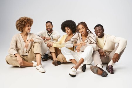 Gruppe fröhlicher afrikanisch-amerikanischer Freunde in lässiger Kleidung auf grauem Hintergrund sitzend, Juni