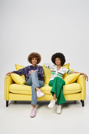 young african american female friends sitting on yellow sofa on grey background, Juneteenth concept
