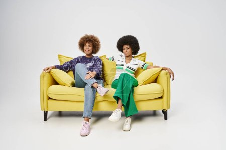 happy african american female friends sitting on yellow sofa on grey background, Juneteenth concept