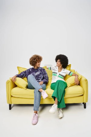 fröhliche afrikanisch-amerikanische Freundinnen sitzen auf gelbem Sofa und plaudern auf grauem Hintergrund
