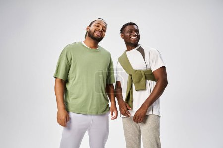 fröhliche afrikanisch-amerikanische männliche Freunde in Freizeitkleidung stehen zusammen auf grauem Hintergrund, Juni