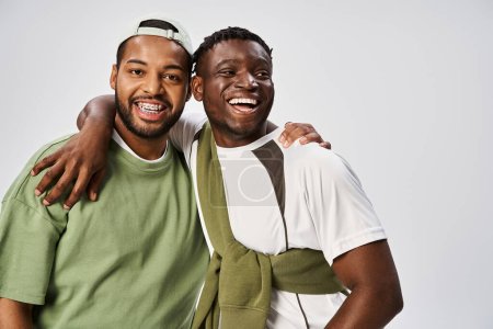 fröhliche afrikanisch-amerikanische männliche Freunde in lässiger Kleidung umarmen sich auf grauem Hintergrund, Juni