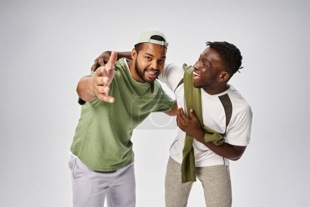 fröhlicher afrikanisch-amerikanischer Mann zeigt mit der Hand in die Nähe eines Freundes auf grauem Hintergrund, Juni