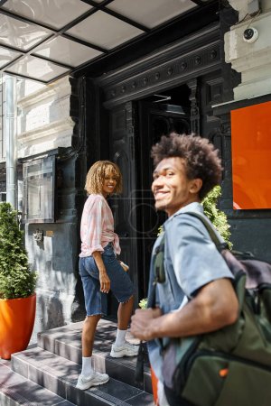 happy african american man near girlfriend entering youth hostel on city street, fun and travel