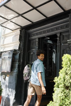 Téléchargez les photos : Étudiant afro-américain joyeux et branché avec sac à dos entrant hôtel de jeunesse dans la rue de la ville - en image libre de droit