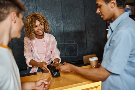 souriant afro-américaine réceptionniste tenant terminal de paiement près des étudiants multiethniques en auberge