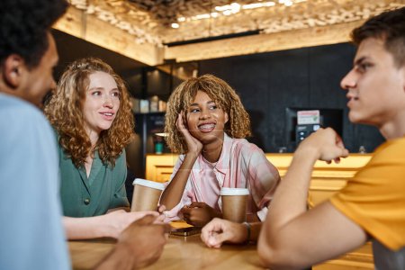 joyful multiethnic students talking near paper cups in lounge cafe of youth hostel, friendship