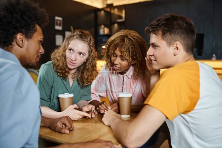 Foto de Jóvenes y elegantes amigos multiétnicos hablando cerca de bebidas para llevar en la cafetería del vestíbulo del albergue de estudiantes - Imagen libre de derechos