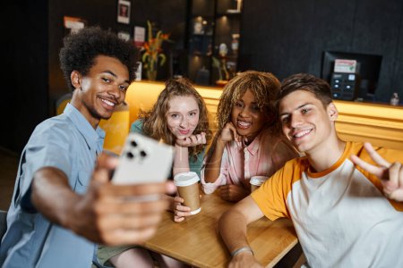 cheerful african american man taking selfie with stylish multiethnic friends in lounge of hostel